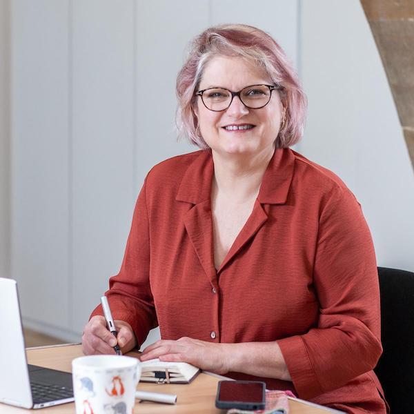 Anne Barrett is sitting writing at a desk, smiling at the camera.