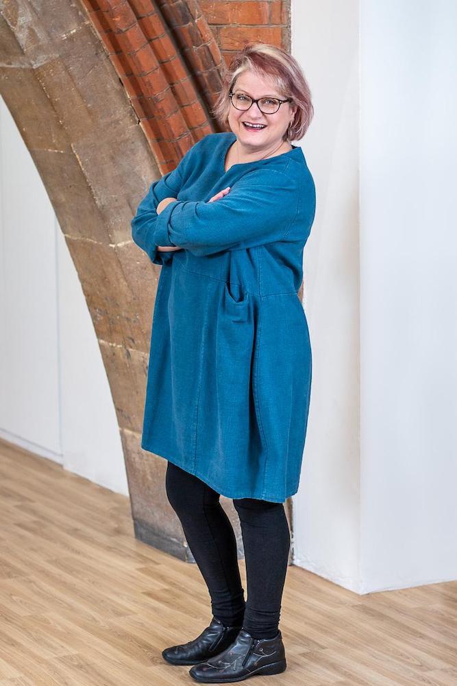 Anne Barret is standing side on with her arms folded, smiling warmly at the camera.